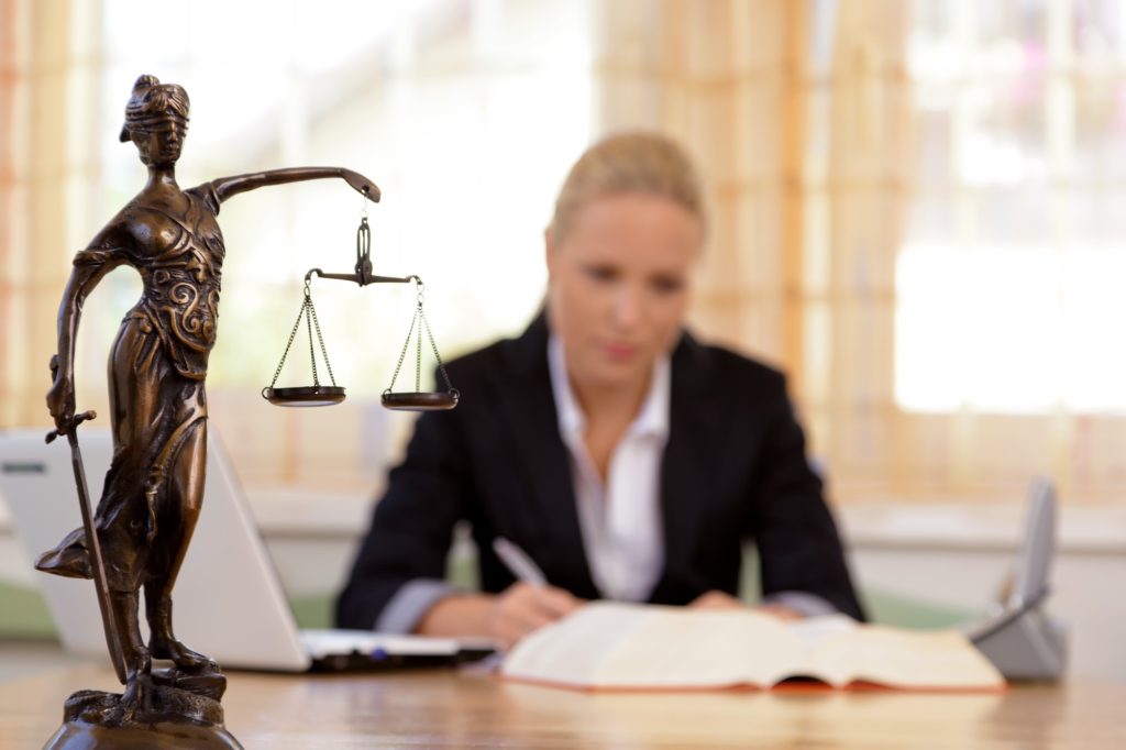 A lawyer sits writing at her desk while the scales of justice are in the foreground