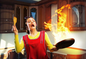 Woman in yellow with Red apron leaning back from a fire in her skillet with a spatula in her other hand standing in a kitchen