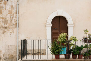 Brown door in white archway in beige wall opening onto a balcony with plants and wrought iron railing