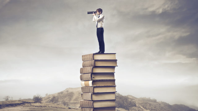 Man with binoculars standing atop a huge stack of books