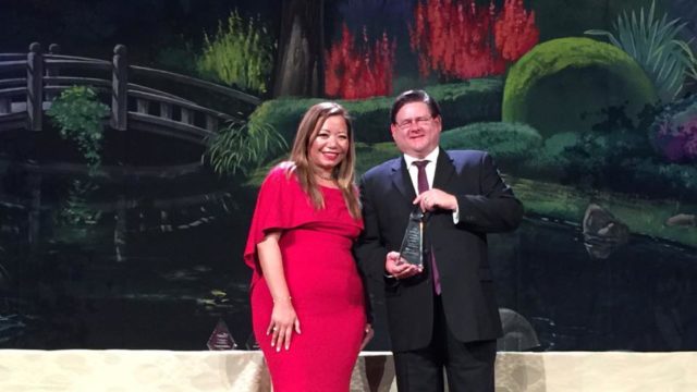 Michael Berg (white male in suite and tie) receiving award on stage with the Executive Director (latinx female in red dress