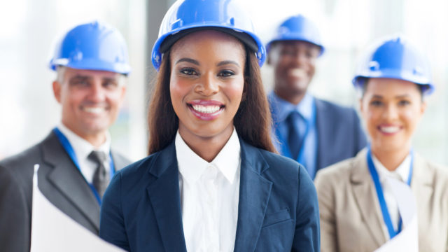 A black woman with long hair, business attire, and a hard hat holding plans with three other people (white male, black male, white female) behind her