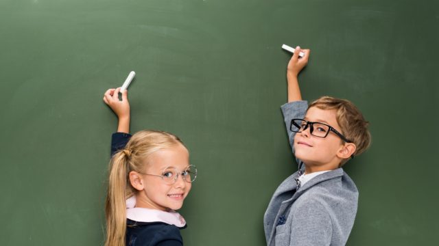 A white little girl, and a white little boy dressed up with glasses on at a chalkboard