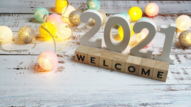 Welcome 2021 displayed as wood blocks and carved numbers on a distressed white wood table with a string of pink, teal, yellow and white round string lights nearby